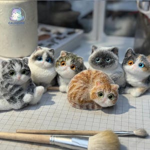 Könnte beinhalten: Eine Sammlung handgefertigter Katzenfiguren in verschiedenen Farben und Mustern. Die Figuren zeigen graue, orangefarbene und weiße Katzen mit detaillierten Merkmalen und ausdrucksstarken Augen. Die Katzen sind auf einer karierten Oberfläche angeordnet, mit Kunstbedarf im Vordergrund.