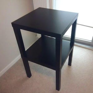 May include: A black square side table with a lower shelf. The table is approximately 24 inches tall and 18 inches wide, with a simple, modern design. The table is made of wood and has a matte finish.