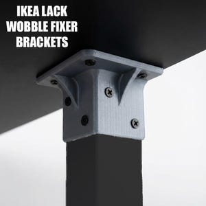 May include: Gray wobble fixer brackets attached to a black table leg. The brackets are secured with black screws. The text "IKEA LACK WOBBLE FIXER BRACKETS" is printed in white.