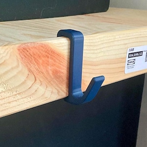 May include: A dark blue hook attached to a light-colored wooden shelf. The hook is designed to hang items and is made of a solid material. The shelf has a label with the text "IVAR" and a product code.