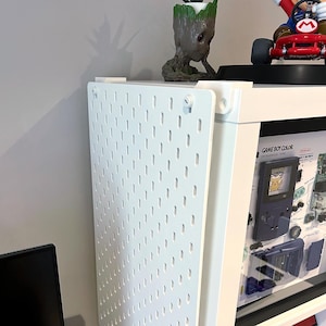 May include: White pegboard with evenly spaced holes, attached to a white frame. A small Groot figurine and a Mario Kart toy sit on top of the frame. A framed Game Boy Color display is visible.