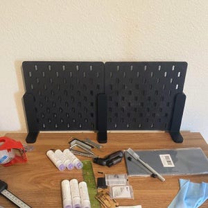 May include: Two black pegboards with numerous holes, standing upright on a wooden surface. Various tools and supplies, including tubes of glue, Allen wrenches, and a ruler, are scattered on the table, suggesting a workspace or craft area.