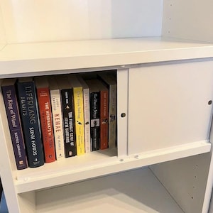 May include: A white bookshelf with a sliding door, partially open to reveal a collection of books. The books have various colored spines and titles, including "The King of Wind" and "Lifelang". The shelf is made of wood and has a clean, modern design.