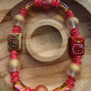May include: A beaded bracelet featuring a mix of pink, gold, and iridescent beads. The bracelet includes gold-toned square charms with a Hello Kitty design. The beads are arranged in a circular pattern, creating a playful and colorful accessory.
