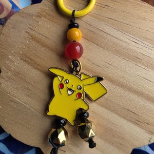 Puede incluir: Un charm de Pikachu amarillo con un cierre amarillo, cuentas negras, naranjas y rojas, y cuentas geométricas doradas. El charm está sujeto a una superficie de madera con una tela estampada azul y blanca en el fondo.