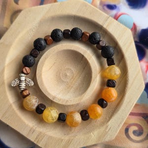 May include: A beaded bracelet featuring black lava rock beads, wooden cubes, and yellow and orange agate beads. A small bee charm is also included. The bracelet is displayed on a wooden octagonal tray.