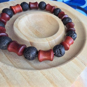 Puede incluir: Una pulsera con cuentas alternas de madera roja oscura y cuentas de roca volcánica negra. La pulsera se exhibe en una bandeja de madera clara, mostrando los colores y texturas naturales de los materiales.