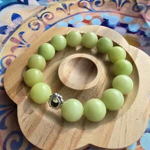 Op de afbeelding: Een lichtgroene kralenarmband met een zilveren accent. De armband is gemaakt van ronde, gladde kralen en wordt gepresenteerd op een houten, bloemvormig dienblad. De achtergrond heeft een kleurrijke stof met patronen.