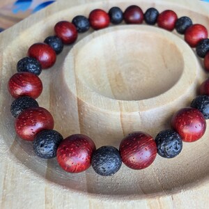 May include: A bracelet made of alternating red wooden beads and black lava beads. The bracelet is presented on a light brown wooden plate. The beads are round and polished. The wooden beads are a deep red.