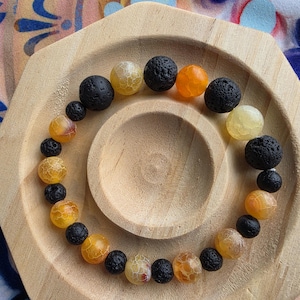 May include: A beaded bracelet featuring a mix of black lava rock and yellow-orange agate beads. The bracelet is displayed on a light wooden, hexagonal-shaped tray.