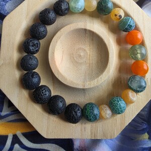 Op de afbeelding: Een kralenarmband met een verscheidenheid aan kleurrijke stenen, waaronder zwarte, groene, oranje en gele, gerangschikt in een cirkelvormig patroon. De armband wordt gepresenteerd op een houten achthoekige tray met een centrale cirkelvormige inkeping. De achtergrond is een patroonstof.