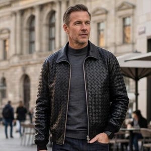 Könnte beinhalten: Schwarze Lederjacke mit gewebtem Muster, Reißverschluss und Kragen. Die Jacke wird über einem grauen Rollkragenpullover und dunkelblauen Jeans getragen. Eine Uhr ist am Handgelenk sichtbar.