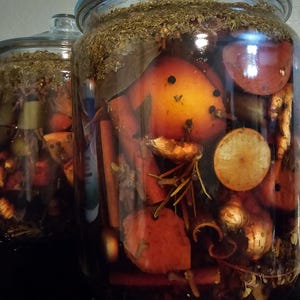 May include: Two large glass jars filled with a dark liquid and various ingredients. The jars contain orange-colored fruits, cinnamon sticks, and herbs. The jars have glass lids and are set against a dark background.