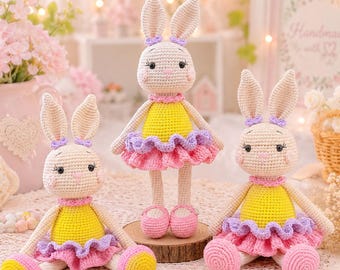Può includere: Tre bambole coniglio all'uncinetto con corpo in filato beige, orecchie lunghe e guance rosa. Ogni coniglio indossa un top giallo, una gonna a balze viola e rosa e scarpe rosa. Il coniglio centrale è posizionato su una base di legno.