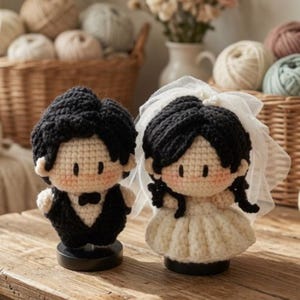 May include: Two crochet dolls representing a wedding couple, one wearing a black suit with a bow tie and the other wearing a white dress with a sheer veil. Both dolls have black yarn hair, light skin tones, and are mounted on small black circular stands.