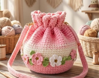 Può includere: Una borsa a secchiello all'uncinetto con coulisse caratterizzata da una sfumatura di filato rosa e bianco. La borsa è decorata con fiori all'uncinetto tridimensionali in bianco, rosa e verde. Include una lunga tracolla all'uncinetto rosa.