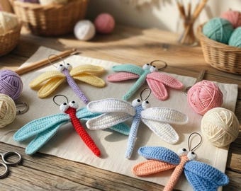 Può includere: Sei ornamenti a forma di libellula realizzati all'uncinetto in vari colori tra cui giallo, rosa, rosso, bianco, blu e arancione, ciascuno con due piccoli occhi bianchi e un anello metallico, disposti su un panno con gomitoli e forbici.