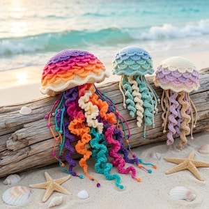 Könnte beinhalten: Drei gehäkelte Quallen mit geschuppten Kappen und spiralförmigen Tentakeln. Die größte ist regenbogenfarben, die mittlere hellblau und die kleinste pastellviolett und cremefarben. Sie liegen auf einem Stück Treibholz an einem Sandstrand.