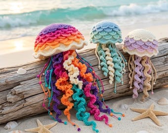 May include: Three crocheted jellyfish toys with scalloped caps and spiral tentacles. The largest is rainbow-colored, the middle is light blue, and the smallest is pastel purple and cream. They are displayed on a piece of driftwood on a sandy beach.