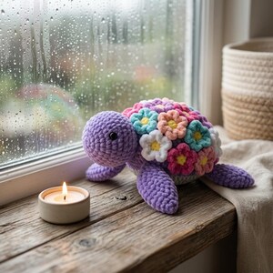 May include: A crocheted turtle plush toy with a purple head and limbs, featuring a shell decorated with numerous small, multi-colored yarn flowers in shades of pink, blue, white, and yellow, positioned next to a small lit candle.