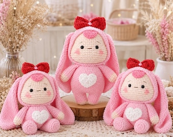 Può includere: Tre bambole lavorate all'uncinetto a mano con corpi rosa, lunghe orecchie da coniglio e un fiocco rosso brillante sulla testa. Ogni bambola ha un viso rosa chiaro, occhi neri ricamati e una toppa a forma di cuore bianco sul petto.