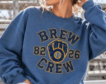 Può includere: Una felpa blu a girocollo con una stampa grafica usurata gialla e blu scuro che recita BREW CREW, con i numeri 82 e 26 su entrambi i lati di un'icona di guantone da baseball contenente una palla al centro del disegno.