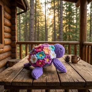 May include: A plush turtle toy crafted from purple yarn, featuring a shell decorated with numerous small, multi-colored crocheted flowers in shades of pink, white, blue, and yellow. The turtle has a rounded head with black bead eyes.