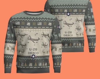 Può includere: Un maglione a maniche lunghe grigio e beige con un motivo ripetuto di aerei U-28 Draco, bandiere americane, piastrine di riconoscimento e alberi di pino. Il design include il testo U-28 DRACO e un emblema a stella su tessuto a maglia.