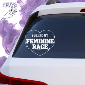 May include: White vinyl car decal with the text "Fueled by Feminine Rage" inside a heart shape. The decal is on the rear window of a white car. The decal also has two starbursts.