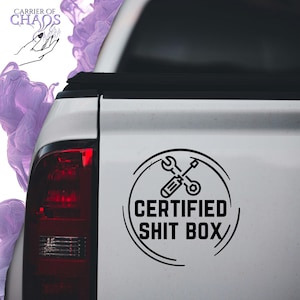 May include: Black and white sticker with the text "CERTIFIED SHIT BOX" inside a circle, featuring crossed wrench and screwdriver icons. The sticker is on the back of a white truck.