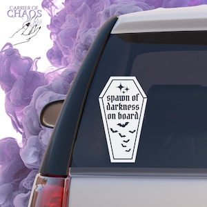 May include: A white vinyl car decal in the shape of a coffin, with the words "spawn of darkness on board" in gothic font. Below the text are several bat silhouettes. The decal is affixed to a car window.