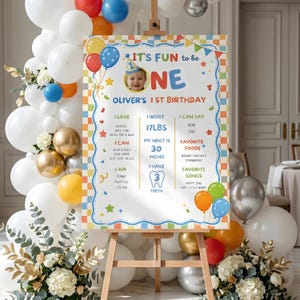 May include: A colorful first birthday sign on an easel, surrounded by balloons and flowers. The sign features a photo of a baby and the text "It's Fun to be ONE." It lists the baby's weight as 17 lbs and height as 30 inches.