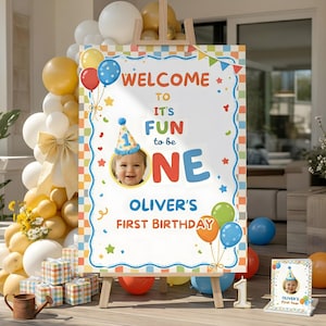 May include: A wooden easel displays a colorful birthday sign that reads "WELCOME TO IT'S FUN to be ONE" with a photo of a baby. Balloons in various colors and a small card with the baby's photo are also present, celebrating a first birthday.