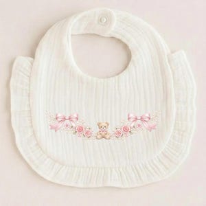 Puede incluir: Babero blanco para bebé con borde con volantes y diseño floral decorativo. El diseño incluye lazos rosas, rosas y un pequeño oso de peluche. El babero tiene un cierre de botón en el cuello.