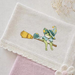 May include: A white knitted baby blanket with a whimsical illustration of a frog wizard riding a broom with a snail. The blanket has a scalloped edge and is folded over a pink blanket.