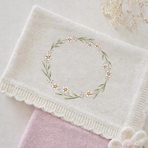 May include: A cream-colored knitted baby blanket with a floral wreath design. The wreath features small white daisies with yellow centers and green leaves. The blanket has a scalloped edge and is folded over a pink blanket.