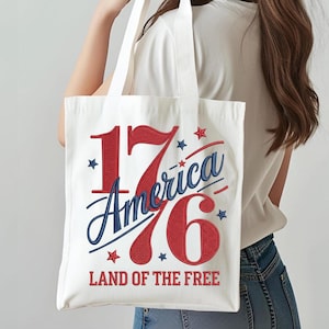 K&ouml;nnte beinhalten: Eine wei&szlig;e Tragetasche mit dem Text "1776 America" in Rot und Blau, mit Sternakzenten. Der Satz "LAND OF THE FREE" ist darunter gedruckt. Die Tasche hat wei&szlig;e Griffe und wird von einer Person getragen.