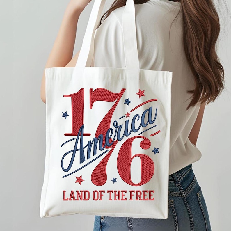 K&ouml;nnte beinhalten: Eine wei&szlig;e Tragetasche mit dem Text "1776 America" in Rot und Blau, mit Sternakzenten. Der Satz "LAND OF THE FREE" ist darunter gedruckt. Die Tasche hat wei&szlig;e Griffe und wird von einer Person getragen.
