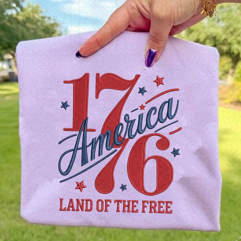 K&ouml;nnte beinhalten: Eine hellviolette Tragetasche mit dem Text "1776 America" in Rot und Blau sowie Sternen. Der Satz "LAND OF THE FREE" ist unten aufgedruckt. Die Tasche wird von einer Hand gehalten.