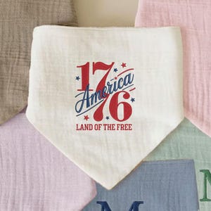K&ouml;nnte beinhalten: Ein wei&szlig;es L&auml;tzchen im Bandana-Stil mit dem Text "1776 America Land of the Free" in Rot und Blau. Das L&auml;tzchen ist von anderen pastellfarbenen Stoffquadraten umgeben.