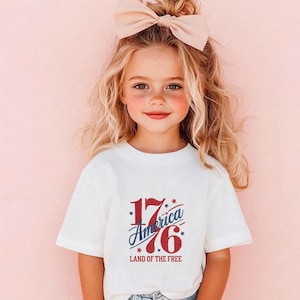 K&ouml;nnte beinhalten: Ein junges Kind tr&auml;gt ein wei&szlig;es T-Shirt mit dem Text "1776 America Land of the Free" in Rot und Blau. Das Kind hat blondes Haar und eine gro&szlig;e Schleife. Der Hintergrund ist einfarbig rosa.