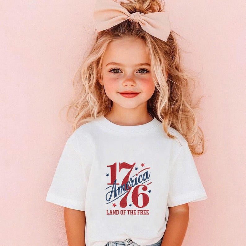 K&ouml;nnte beinhalten: Ein junges Kind tr&auml;gt ein wei&szlig;es T-Shirt mit dem Text "1776 America Land of the Free" in Rot und Blau. Das Kind hat blondes Haar und eine gro&szlig;e Schleife. Der Hintergrund ist einfarbig rosa.