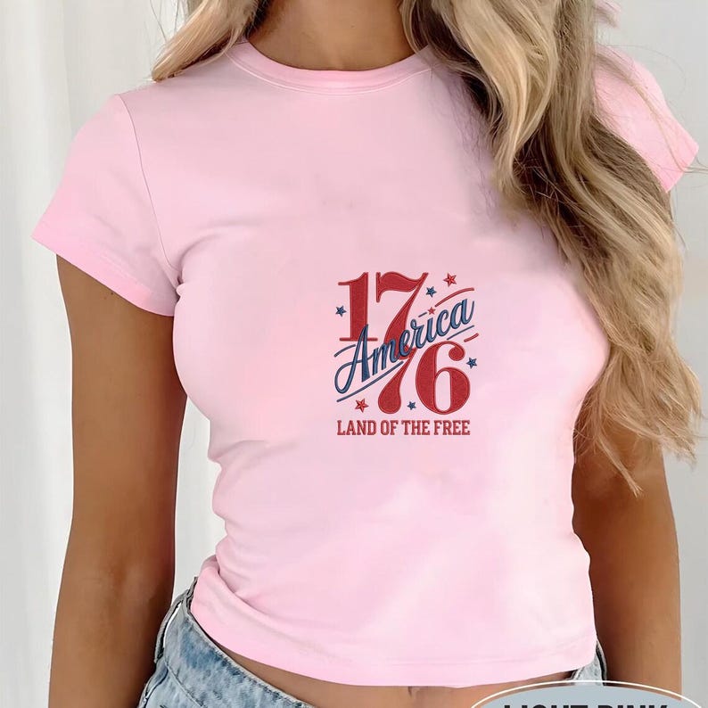 K&ouml;nnte beinhalten: Hellrosa T-Shirt mit Rundhalsausschnitt und kurzen &Auml;rmeln. Auf der Vorderseite steht der Text "1776 America Land of the Free" in Rot, Wei&szlig; und Blau. Das Design umfasst Sterne und eine stilisierte Schriftart.