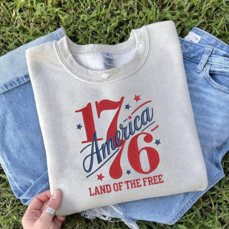 K&ouml;nnte beinhalten: Ein hellgraues Sweatshirt mit dem gestickten Text "1776 America Land of the Free" in Rot und Blau. Das Sweatshirt liegt gefaltet auf einer blauen Jeans, mit gr&uuml;nem Gras im Hintergrund. Das Design feiert die amerikanische Unabh&auml;ngigkeit.