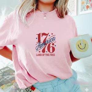 K&ouml;nnte beinhalten: Ein hellrosa Sweatshirt mit dem Text "1776 America Land of the Free" in Rot und Blau. Eine wei&szlig;e Tasse mit einem gelben Smiley wird in der rechten Hand gehalten.