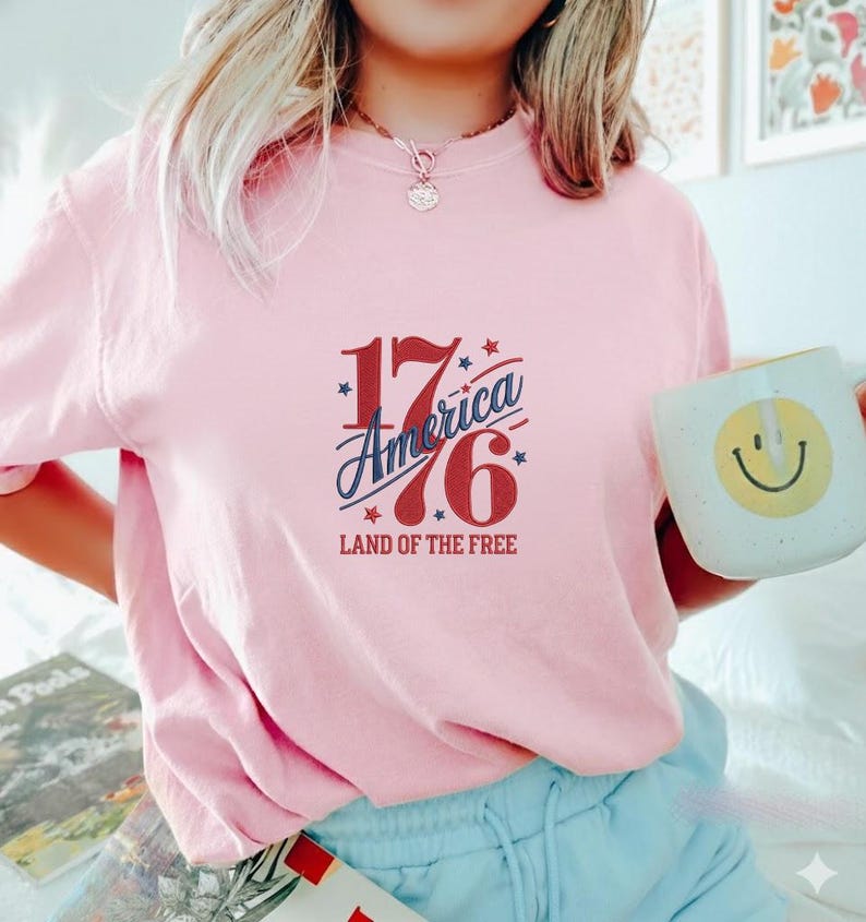 K&ouml;nnte beinhalten: Ein hellrosa Sweatshirt mit dem Text "1776 America Land of the Free" in Rot und Blau. Eine wei&szlig;e Tasse mit einem gelben Smiley wird in der rechten Hand gehalten.