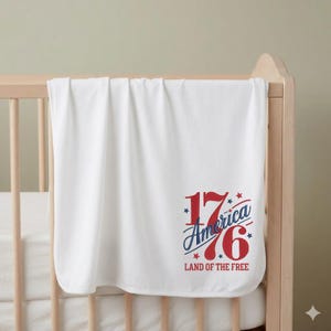 K&ouml;nnte beinhalten: Eine wei&szlig;e Babydecke mit dem Text "1776 America Land of the Free" in roter und blauer Schrift, mit Sternakzenten. Die Decke ist &uuml;ber ein Holzbettchen drapiert. Die Decke wirkt weich und gefaltet.