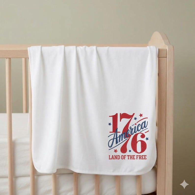K&ouml;nnte beinhalten: Eine wei&szlig;e Babydecke mit dem Text "1776 America Land of the Free" in roter und blauer Schrift, mit Sternakzenten. Die Decke ist &uuml;ber ein Holzbettchen drapiert. Die Decke wirkt weich und gefaltet.