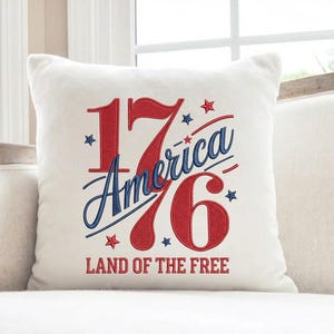 K&ouml;nnte beinhalten: Ein quadratisches wei&szlig;es Kissen mit dem Text "1776 America Land of the Free" in Rot und Blau. Die Zahlen und der Text sind gestickt, mit Sternakzenten.