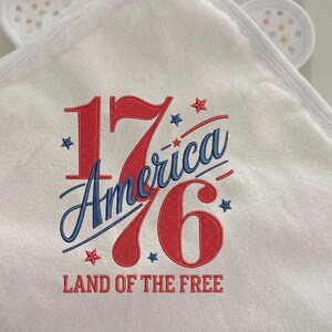 K&ouml;nnte beinhalten: Wei&szlig;es Kapuzenhandtuch f&uuml;r Babys mit gesticktem Text "1776 America" in Rot und Blau, mit Sternen. Der Text "Land of the Free" ist ebenfalls rot gestickt. Das Handtuch hat kleine, wei&szlig;e Ohren mit bunten Punkten.